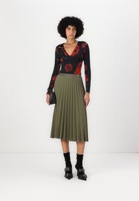 Body nero con stampa di rose rosse, abbinato a una gonna midi plissettata verde oliva. La modella indossa calzini neri alla caviglia e scarpe con punta e tacchi con fibbie.