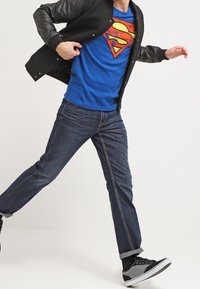 Niebieska koszulka z logo Supermana w czerwonym i żółtym kolorze, czarna kurtka bomberka, ciemne jeansy z denimu i czarne trampki z białą podeszwą.