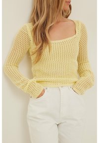 Pull jaune tricoté avec un col carré et des manches longues. Il présente une texture à maille ouverte ainsi que des poignets et un ourlet côtelés. Associé à un jean blanc.