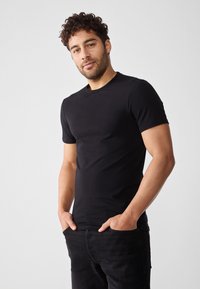 Svart bomulls t-skjorte med korte ermer, rund hals, tettsittende design, brukt sammen med mørke jeans, vist mot en lys bakgrunn.