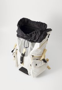 Reppu mustalla narupäällä, valkoisella rungolla, verkkopaneelilla ja keltaisilla hihnoilla. Sisältää useita vetoketjullisia taskuja sivuilla ja edessä.