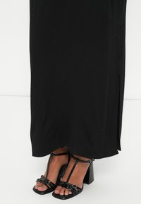 Rochie maxi neagră cu puncte texturate subtile, având o fendă pe lateral. Împerecheată cu sandale negre cu toc înalt, cu designe cu curele și catarame decorative.