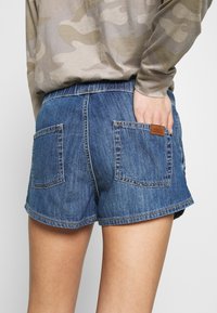 Pantalones cortos de mezclilla en azul medio, con un corte relajado, dos bolsillos traseros y una etiqueta con el logo marrón cerca de la cinturilla. La tela tiene una textura suave.