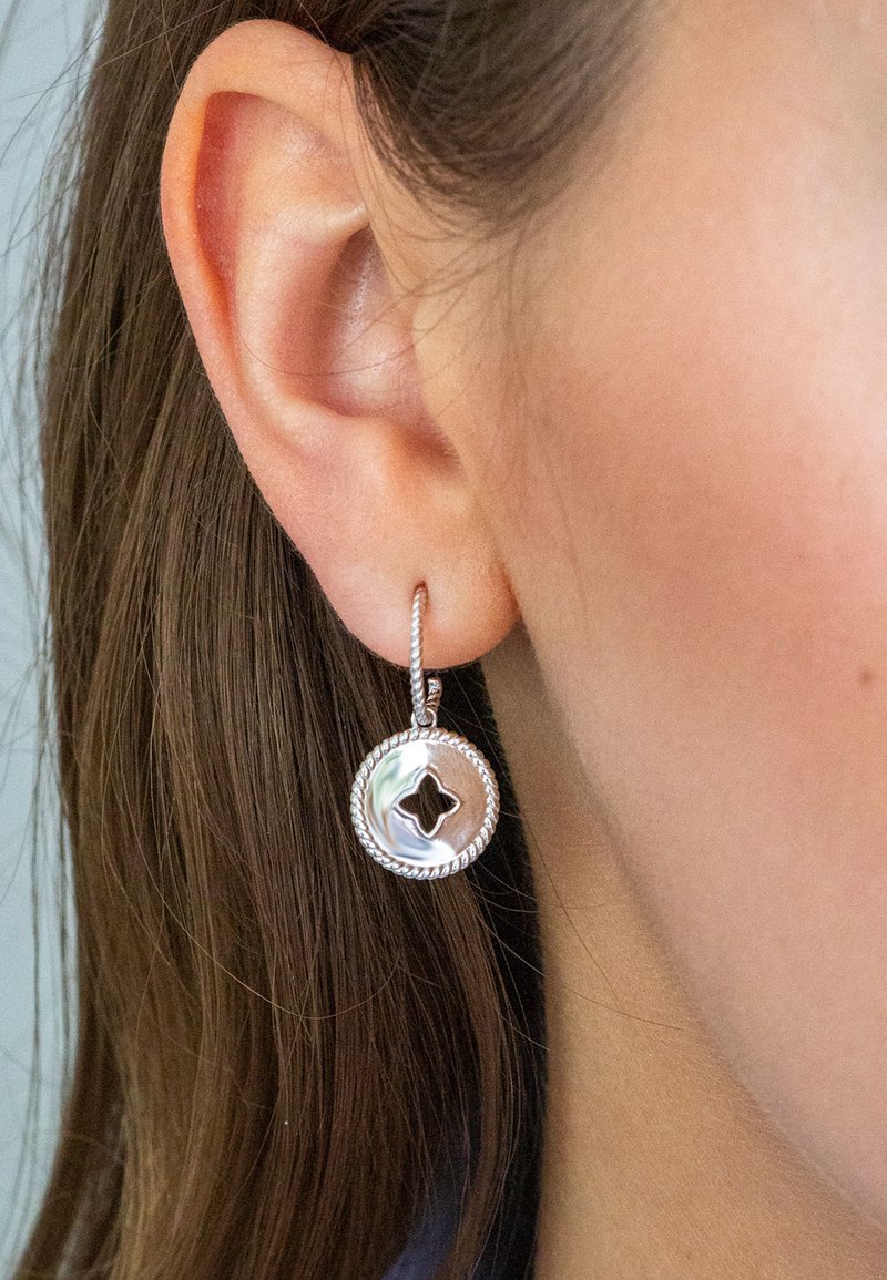 Silberne Creolen mit geflochtenem Rand und einer ausgeschnittenen Sternform in der Mitte, die an einem Ohr einer Person vor einem neutralen Hintergrund präsentiert werden.