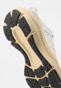Σόλα αθλητικού παπουτσιού σε μπεζ και μαύρο χρώμα με υφή από καουτσούκ, που δείχνει υποστήριξη καμάρας και σχέδιο πέλματος για πρόσφυση, με φόντο λευκό υλικό επάνω μέρους.
