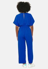 Modrá kombinéza s krátkymi rukávmi, elastickým pásom, detailom v tvare kľúčovej dierky na chrbte a širokými nohavicami, kombinovaná s bielymi teniskami s farebnými akcentmi.