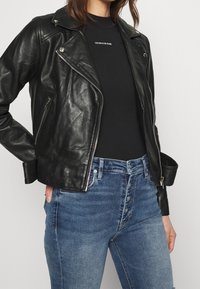 Mujer vestida con chaqueta de cuero negra de moto sobre una blusa negra de Calvin Klein y jeans azul de tiro alto con botón, con la mano en el bolsillo.