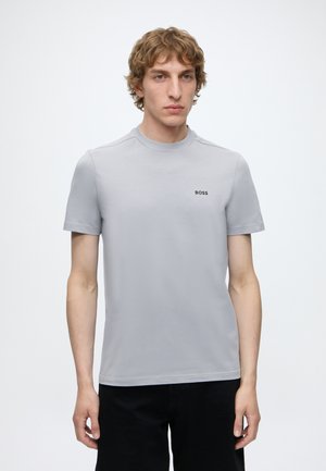 Jeune homme aux cheveux blonds bouclés portant un T-shirt gris clair à manches courtes avec le logo "BOSS" sur la poitrine et un pantalon noir.