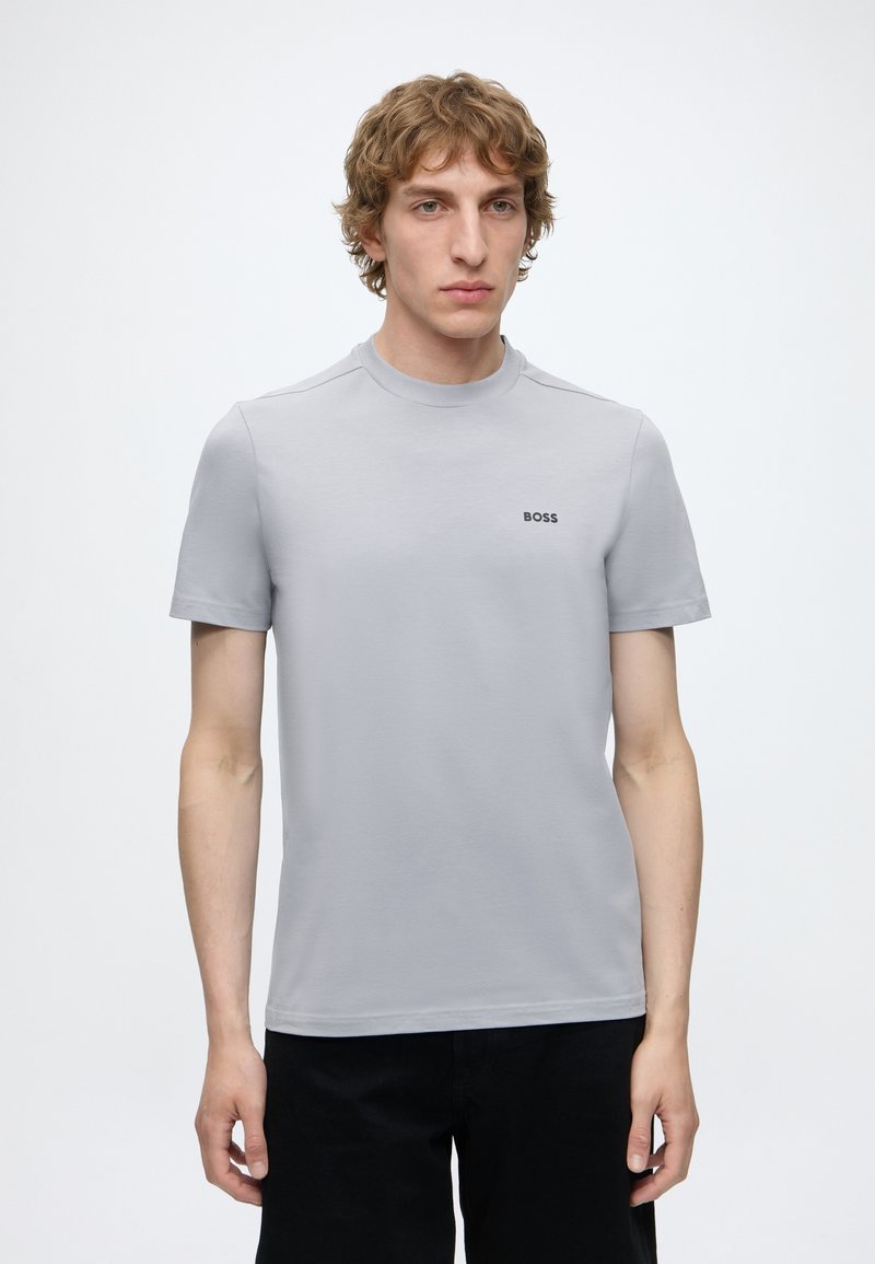 Jeune homme aux cheveux blonds bouclés portant un T-shirt gris clair à manches courtes avec le logo "BOSS" sur la poitrine et un pantalon noir.