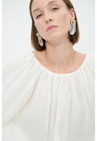 Weiße, strukturierte Bluse mit einem gerafften, runden Ausschnitt. Das Modell trägt große, durchsichtige Ohrringe mit goldenen Akzenten. Weicher, fließender Stoff.