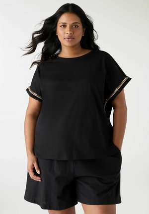 Donna con capelli lunghi e neri, che indossa una maglietta nera a maniche corte con rifiniture dorate e pantaloncini neri, in piedi davanti a uno sfondo chiaro e uniforme.
