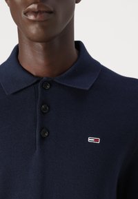 Tamsiai mėlynas megztinis polo su trimis sagomis, koloritu ir mažai siuvinėta logotipu raudona, balta ir mėlyna spalvomis.