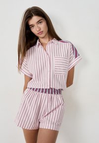 Jeune femme portant une chemise à rayures verticales roses et violettes à manches courtes et un short élastique assorti, se tenant devant un fond blanc.