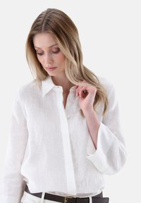 White, long-sleeve linen shirt with a button-down front, a structured collar, and slightly flared sleeves. Worn with a brown belt.