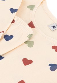 Top en coton beige avec un motif de cœurs colorés en vert, rouge et bleu. Texture douce avec un col rond et un design ludique.