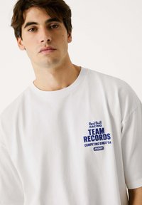 Witte katoenen t-shirt met korte mouwen, voorzien van blauw geborduurde tekst: "Red Bull Racing TEAM RECORDS COMPETING SINCE '04" op de borst.