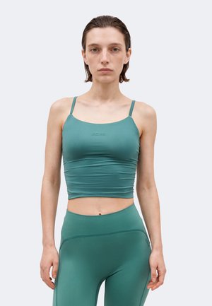 Γυναίκα με στενό τοπ σε πετρόλ χρώμα με λεπτές τιράντες και κολάν με ψηλή μέση, στέκεται μπροστά σε απλό ανοιχτό φόντο.