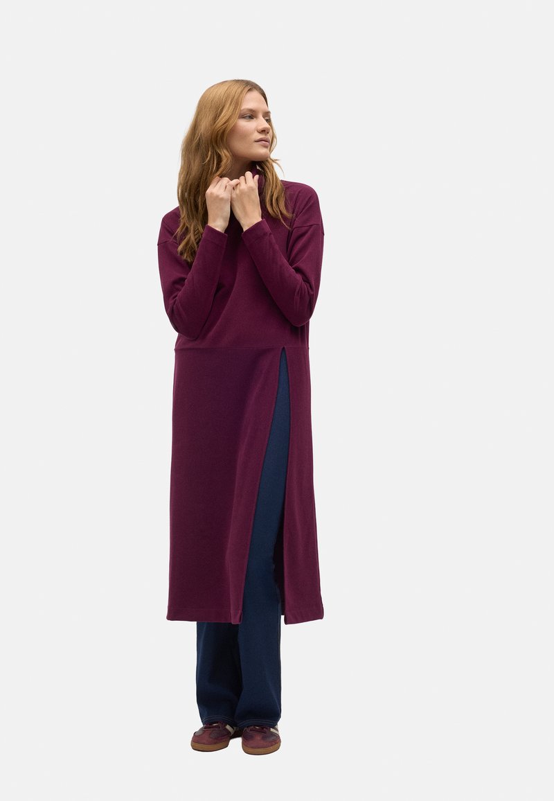 Vestido largo burdeos de manga larga con una abertura lateral, llevado sobre pantalones anchos azul marino, presenta un ajuste holgado y una textura suave, combinado con zapatos marrones.