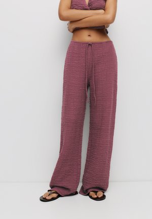 Pflaumenfarbene, gerüschte Stoffhose mit einem Kordelzugbund; weit geschnittenes Design; glatte Textur; kombiniert mit flachen schwarzen Sandalen.