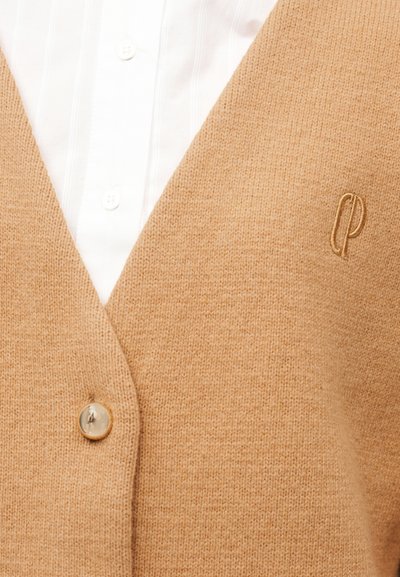 Μπεζ πλεκτή ζακέτα από μαλλί με λαιμόκοψη V, κλείσιμο με ένα κουμπί, υφή πλέξης και κεντημένο λογότυπο στην κάτω γωνία. Κάτω φαίνεται λευκό πουκάμισο με γιακά.