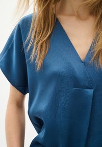 Bluză albastră, lejeră, cu un material texturat, decolteu în V și mâneci scurte. Include o mică buzunar în față.