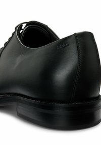 Scarpa da uomo in pelle nera con superficie liscia e forma arrotondata. Presenta un logo discreto vicino al tallone e una suola nera a strati.