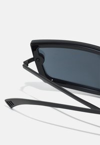 Lunettes de soleil noires avec une monture rectangulaire, des verres foncés et des branches élégantes dotées d'accents de style ventilation pour un design moderne.