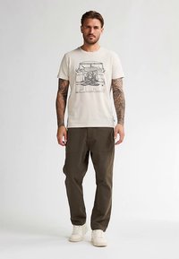 Vaaleanbeige puuvillainen T-paita, jossa on klassisen auton kuva. Yhdistetty oliivinvihreisiin housuihin, jotka on valmistettu kestävästä kankaasta, ja valkoisiin tennareihin.