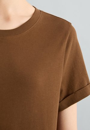 Zbliżenie osoby noszącej brązową koszulkę z krótkim rękawem i okrągłym dekoltem z detalem złożonego rękawa na jasnoszarym tle.