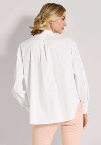 Frau mit blonden Haaren, die zur Seite schaut, trägt ein lockeres weißes Langarmshirt und helle rosa Hosen vor einem schlichten Hintergrund.
