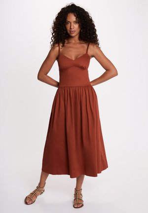 Vrouw met krullend haar, staand met handen op de heupen, draagt een roestkleurige mouwloze midi-jurk en gespen met studs.