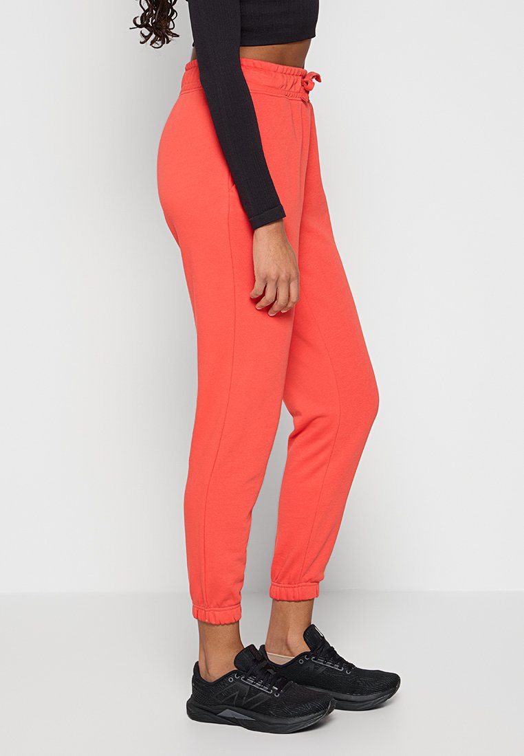 Frau in korallenfarbenen Jogginghosen, schwarzem langärmeligen Crop-Top und schwarzen Sportschuhen, die vor einem schlichten hellen Hintergrund steht.