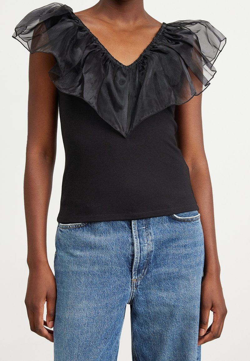 Svart ärmlös topp med genomskinlig volang V-ringad krage, bärs med blå högmidjade jeans av en stående person.