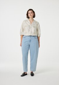 Cremfarbene Bluse mit Sonnenmustern, lockere Passform und halblangen Ärmeln, kombiniert mit hellblauen high-waisted Jeans und schwarzen Slippern mit goldenen Akzenten.