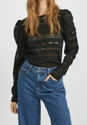 Kvinde iført en sort langærmet blonde top med puffede skuldre og højtaljede blå jeans, hånd afslappet i lommen.