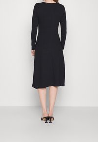 Robe midi noire avec manches longues, corsage ajusté et jupe évasée. Texture lisse, couture arrière subtile, associée à des escarpins noirs à bout pointu.