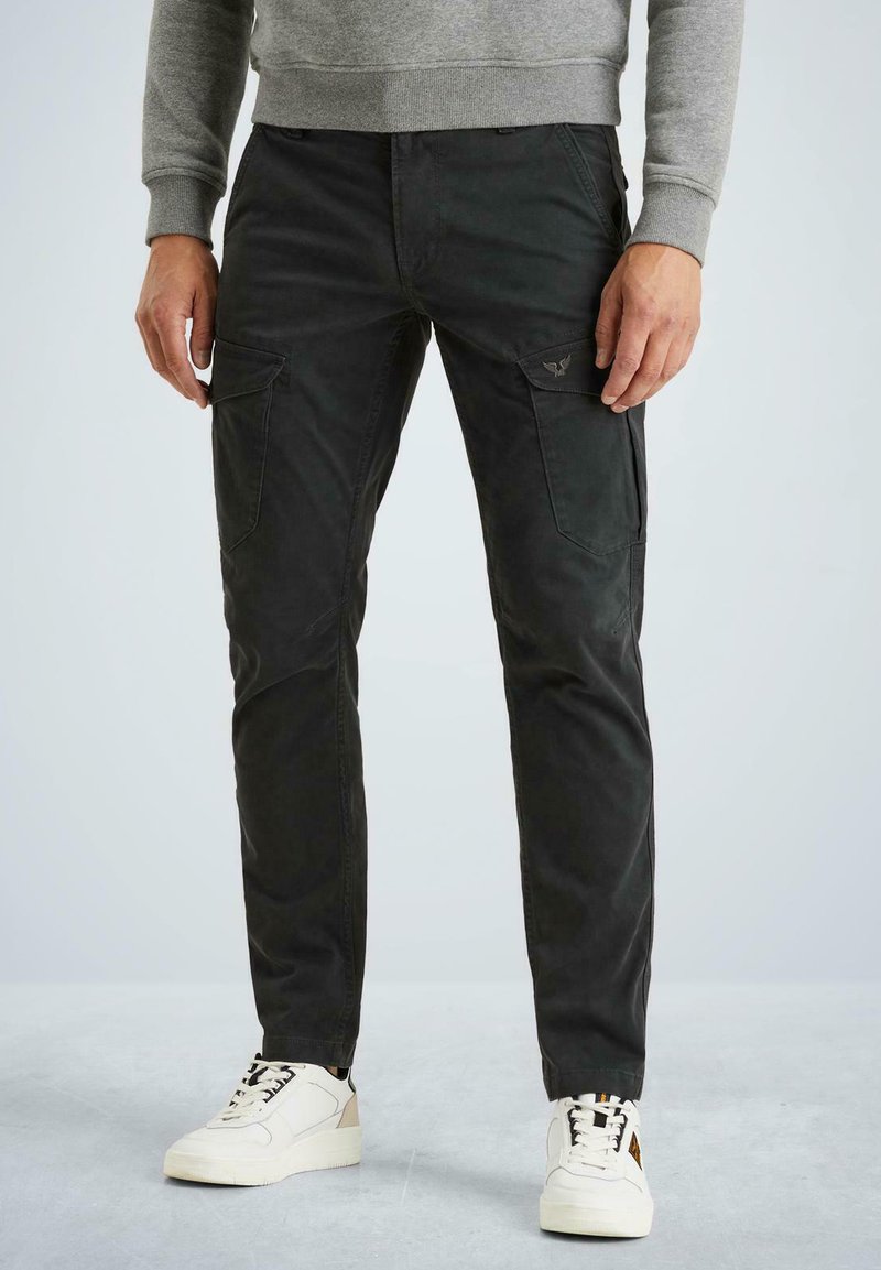 Muž v černých kalhotách s bočními kapsami a bílých teniskách na jednobarevném světlém pozadí.