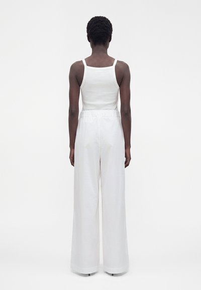 Personne aux cheveux courts et bouclés, portant un haut blanc sans manches et un pantalon blanc taille haute à jambes larges, debout de dos.