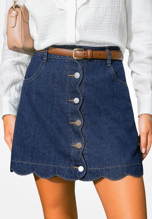 Falda vaquera azul oscuro con dobladillo ondulado y botones plateados en el frente, combinada con un cinturón marrón y una blusa blanca con textura.
