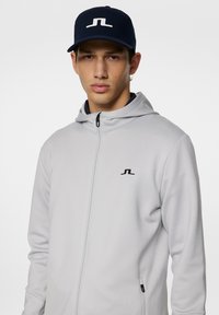 Grauer Reißverschluss-Hoodie mit Kapuze, ausgestattet mit einem schwarzen Logo auf der linken Brust. Kombiniert mit einer marineblauen Kappe, die ein weißes Logo zeigt.