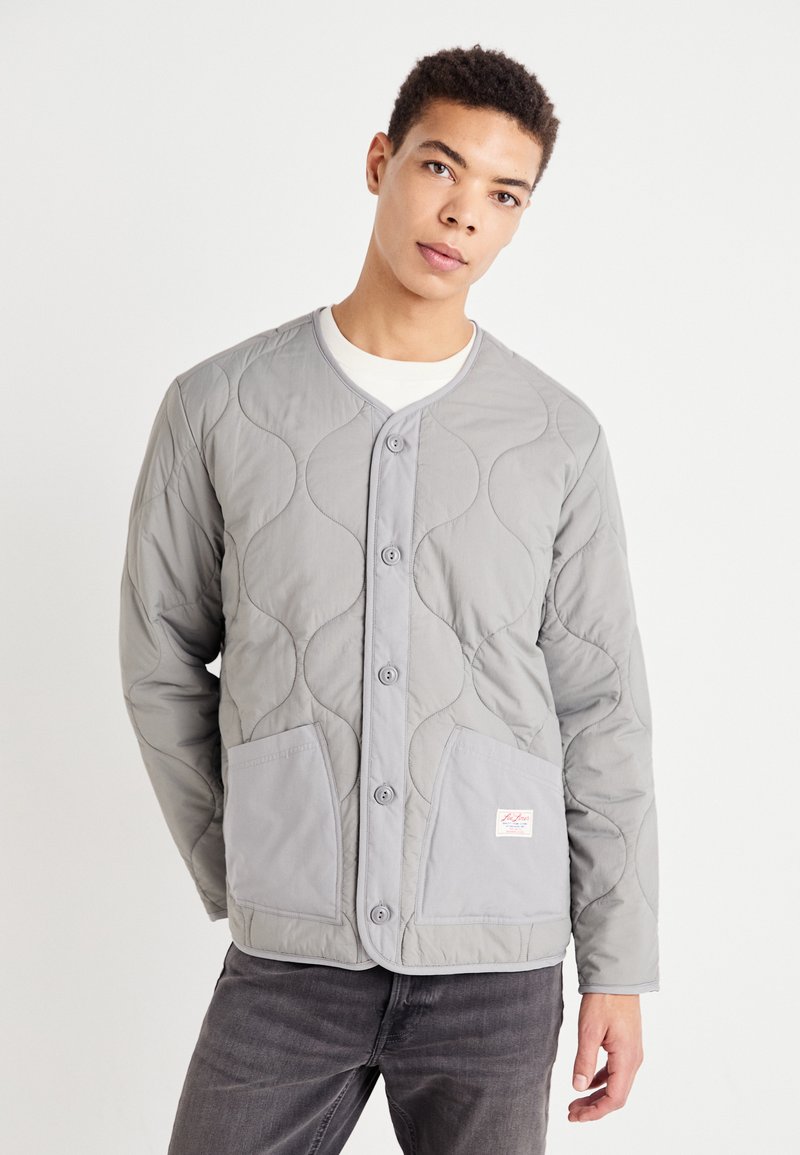 Veste grise matelassée avec un col rond, fermeture à boutons, deux poches avant et un motif en vagues subtile sur le tissu. Design léger et décontracté.