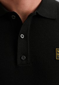 Zwarte polo met een gestructureerd breipatroon, voorzien van een kraag, twee zwarte knopen en een klein geel logo-opdruk op de borst.