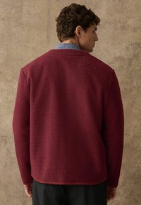 Burgunderfarbene Fleecejacke mit einer strukturierten Oberfläche, lockerer Passform und Rückendetails. Kombiniert mit einem blauen Jeanshemd darunter.