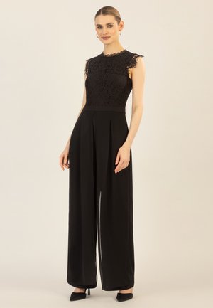 Junge Frau trägt einen ärmellosen schwarzen Spitzen-Overall mit weitem Bein und schwarzen Absätzen, steht vor einem einfarbigen Hintergrund.