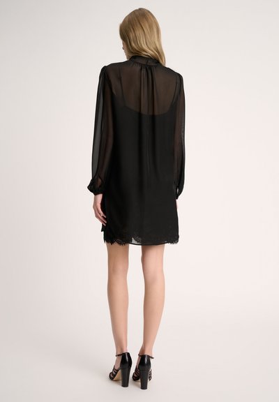 Vestido negro transparente con mangas largas, con un borde de encaje en el dobladillo. Presenta un cuello alto y un corte ligeramente suelto, acompañado de sandalias negras de tacón.