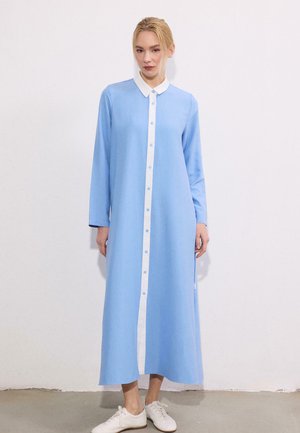 Donna in piedi contro una parete bianca, indossa un lungo vestito azzurro chiaro con bottoni, colletto bianco e sneaker bianche.