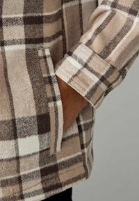 Beżowa i brązowa koszula flanelowa w kratę, z mankietami na guziki i kieszonką boczną. Teksturowana tkanina o miękkim, przytulnym wyglądzie.