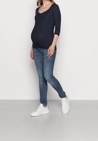 Marineblaues Oberteil mit Dreiviertelärmeln und V-Ausschnitt, kombiniert mit hellblauen Skinny-Jeans und weißen Sneakers, vor einem schlichten Hintergrund.