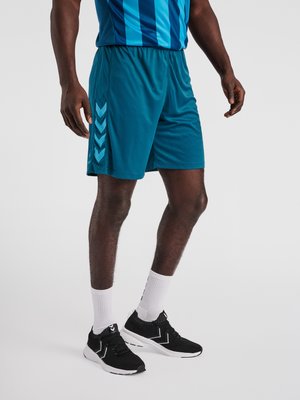 Man draagt blauwe sportshorts en een gestreept shirt, witte sokken en zwarte sportschoenen op een effen grijze achtergrond.