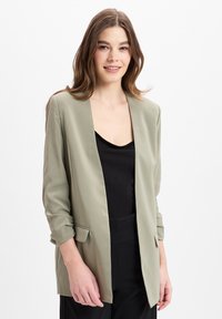 Lichtgroene blazer, ontwerp met open voorkant, lange mouwen met opgerolde manchetten, gemaakt van soepel stof, gecombineerd met een zwarte top eronder.
